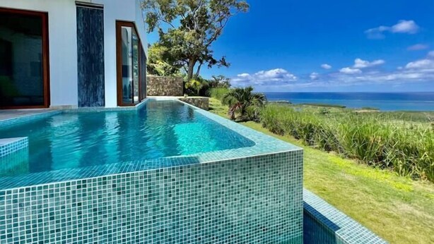 A modern pool with clear blue water, surrounded by lush greenery and a view of the ocean.