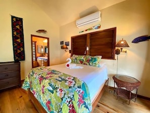 A bedroom with a bed, a dresser, an air conditioning unit, a lamp, and a chair.