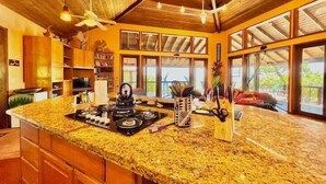 A kitchen with a granite countertop, wooden cabinets, and a large island. There are multiple windows providing natural light and views of the outdoors.