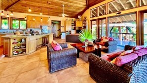 A spacious living area with wicker furniture, a kitchen, and large windows offering a view of the outdoors.