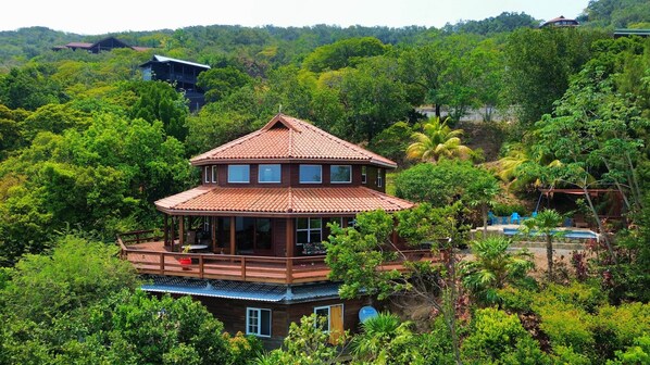 A two-story wooden house with a tiled roof, surrounded by lush greenery and a pool. - Villa con Vista @ Calabash Bight, Roatan. (Oakridge)