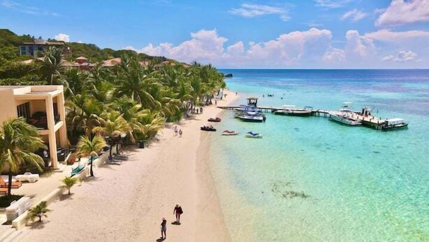 Una playa con agua turquesa cristalina, un muelle con barcos y un edificio con balcones.
