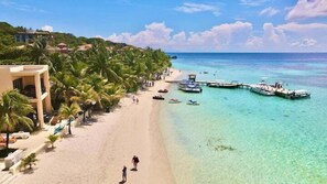 A beach with clear turquoise water, a pier with boats, and a building with balconies. - Coral Sand By The Beach @ West Bay, Roatan. (West Bay)