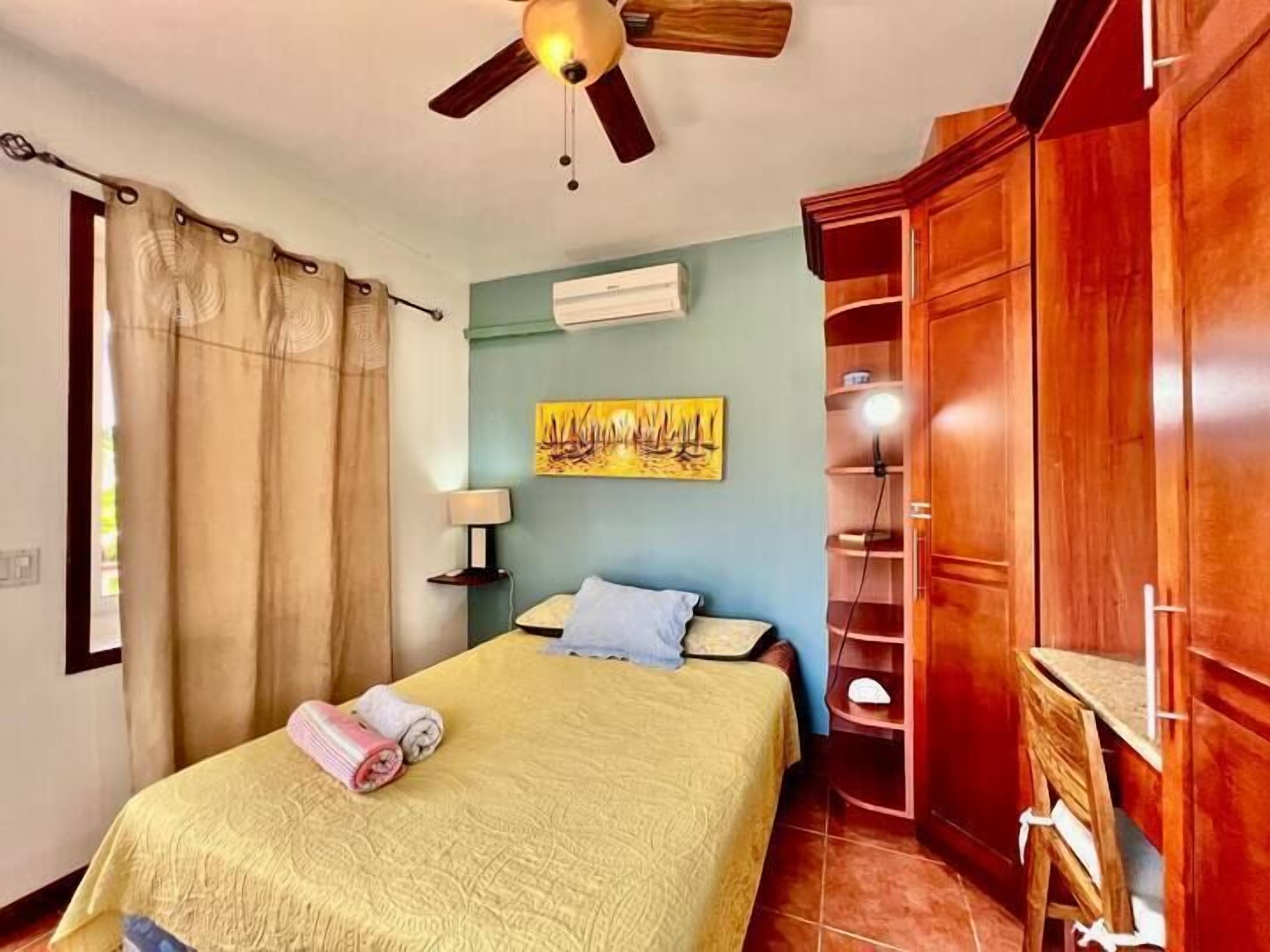 A bedroom with a bed, a ceiling fan, a painting, and a wooden wardrobe.