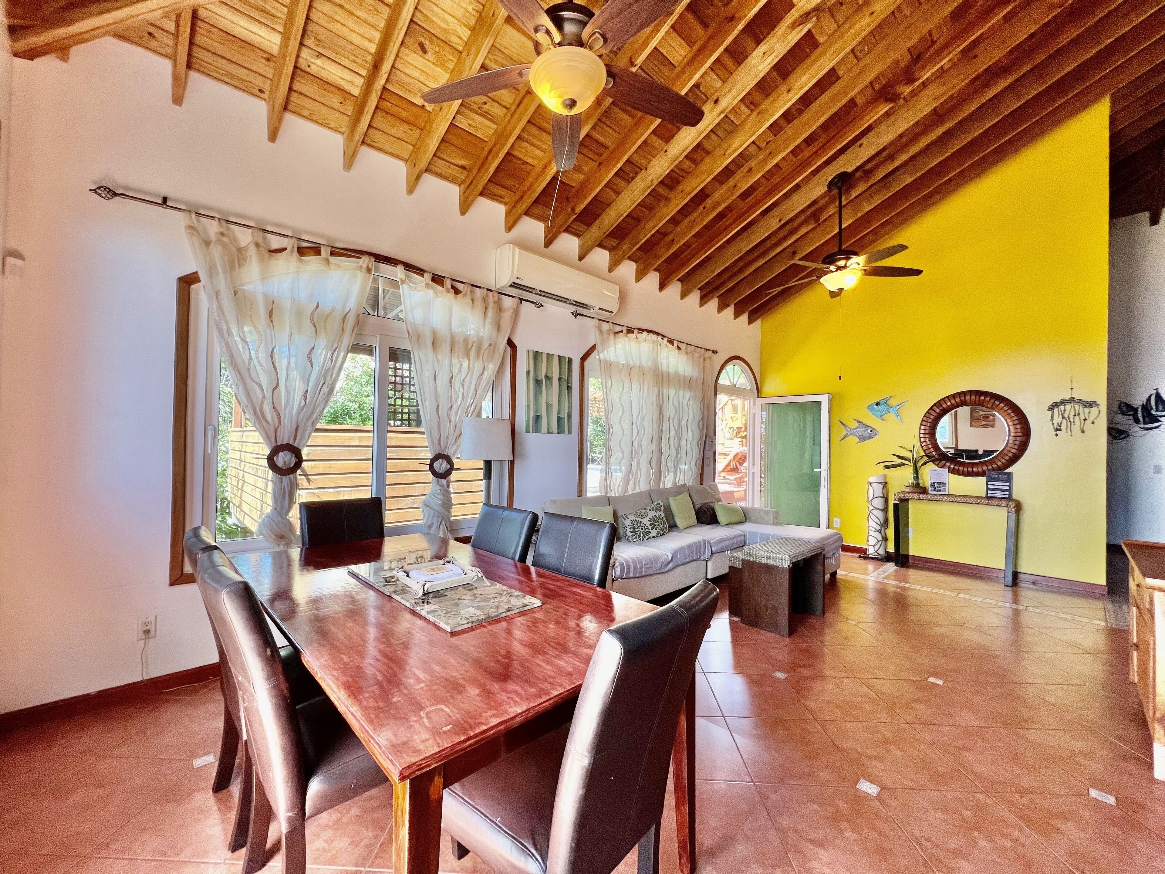 A dining area with a wooden ceiling, a dining table with chairs, a sofa, and a yellow wall with a mirror and wall art.