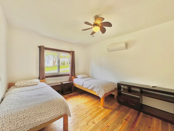 Ein kleiner, gemütlicher Raum mit zwei Einzelbetten, Holzboden, Deckenventilator, Fenster mit Vorhängen und Klimaanlage.