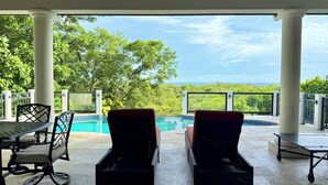 Ett takförsett utomhusområde med en pool, två fåtöljer i rotting, ett bord och utsikt över havet.