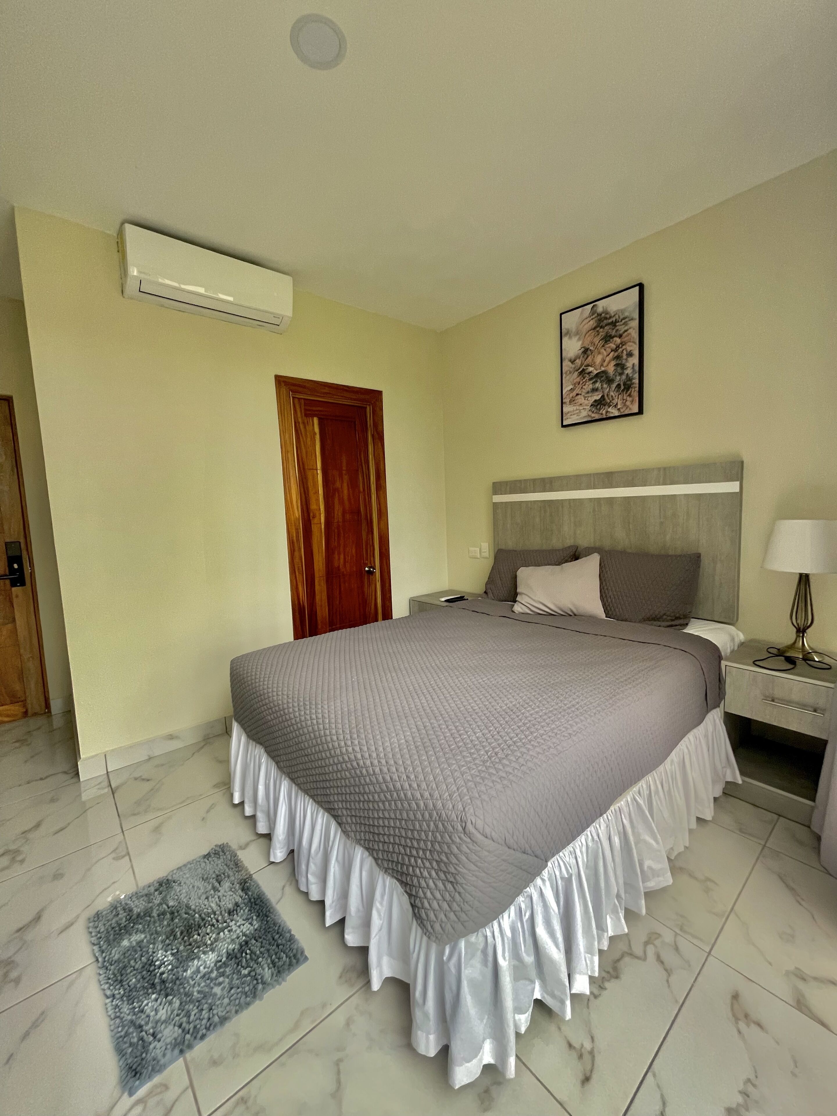 A neatly arranged bedroom with a large bed, a bedside table with a lamp, a wall-mounted air conditioner, and a door.
