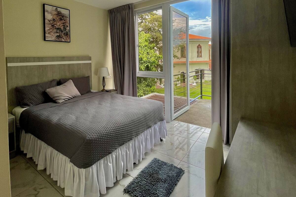 Un dormitorio con cama, mesita de noche, lámpara, ventana con cortinas y vista a un edificio.