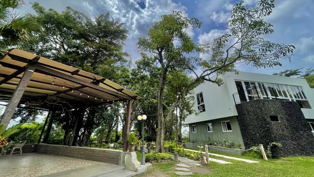 Un espacio de estar al aire libre cubierto con una pérgola, un sendero que lleva a un edificio de fachada de piedra y abundante vegetación alrededor de la propiedad.