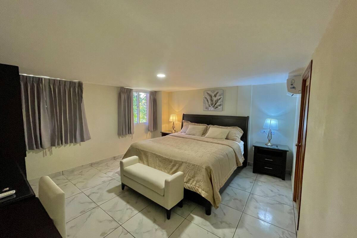 Un dormitorio con cama, mesitas de noche, una silla, una ventana con cortinas y una obra de arte en la pared.