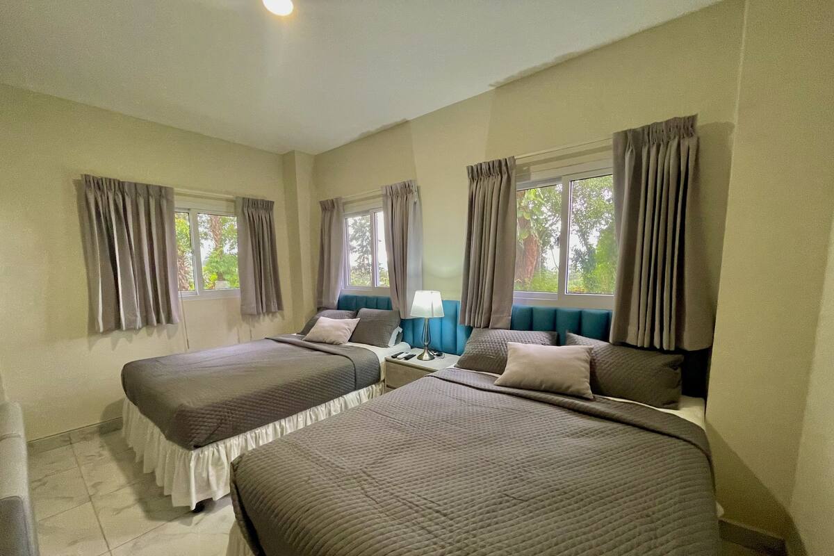 Habitación de hotel con dos camas, una mesita de noche, una lámpara y ventanas con cortinas.