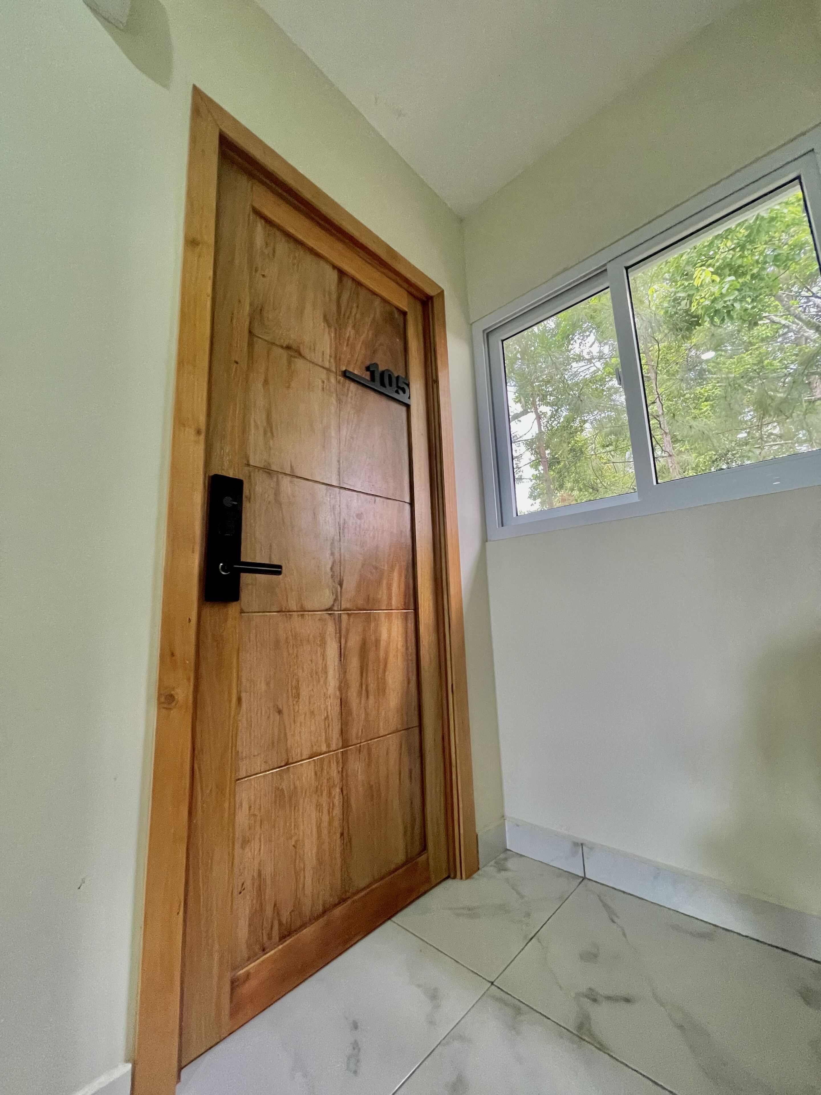 A wooden door with a number plate, a window with a view of trees, and a tiled floor.