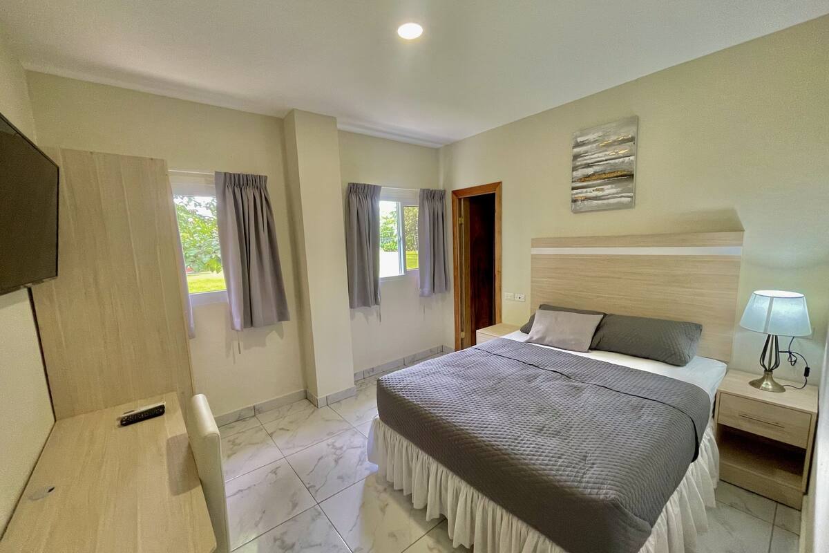 Un dormitorio con cama, mesita de noche, lámpara y televisión a la izquierda.