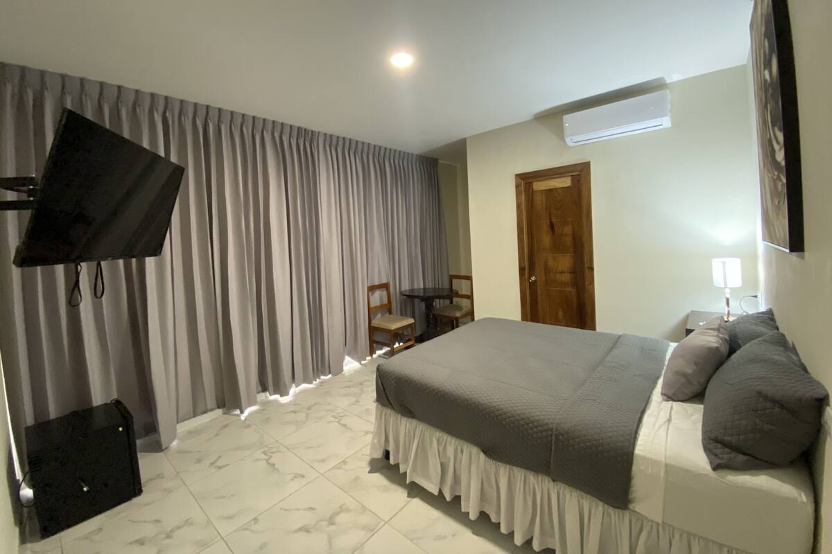 Un dormitorio con cama, cortinas, televisor, una silla, una mesa y aire acondicionado.