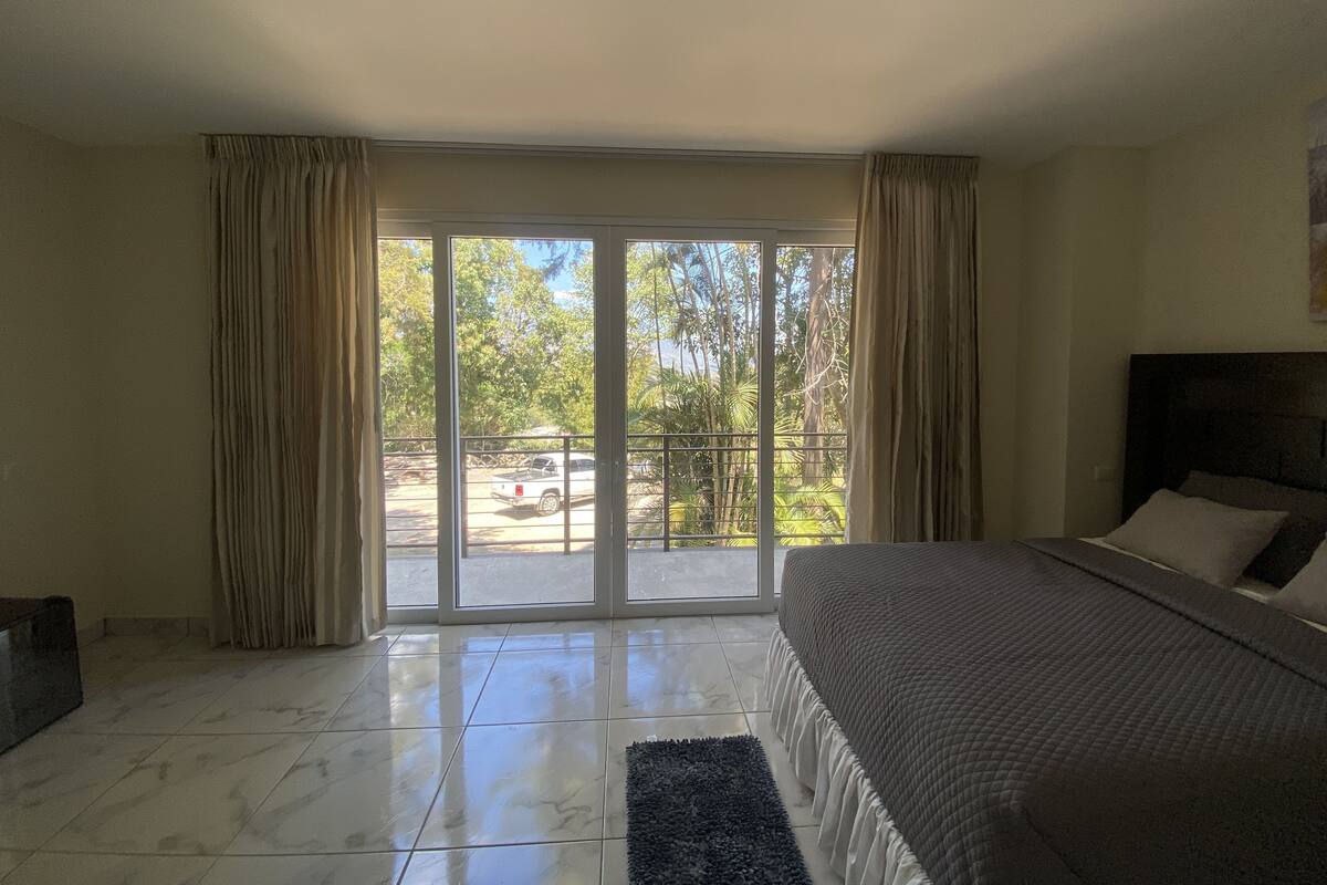 Un dormitorio con una cama grande, un ventanal corredizo que da a un balcón y suelo de baldosas.