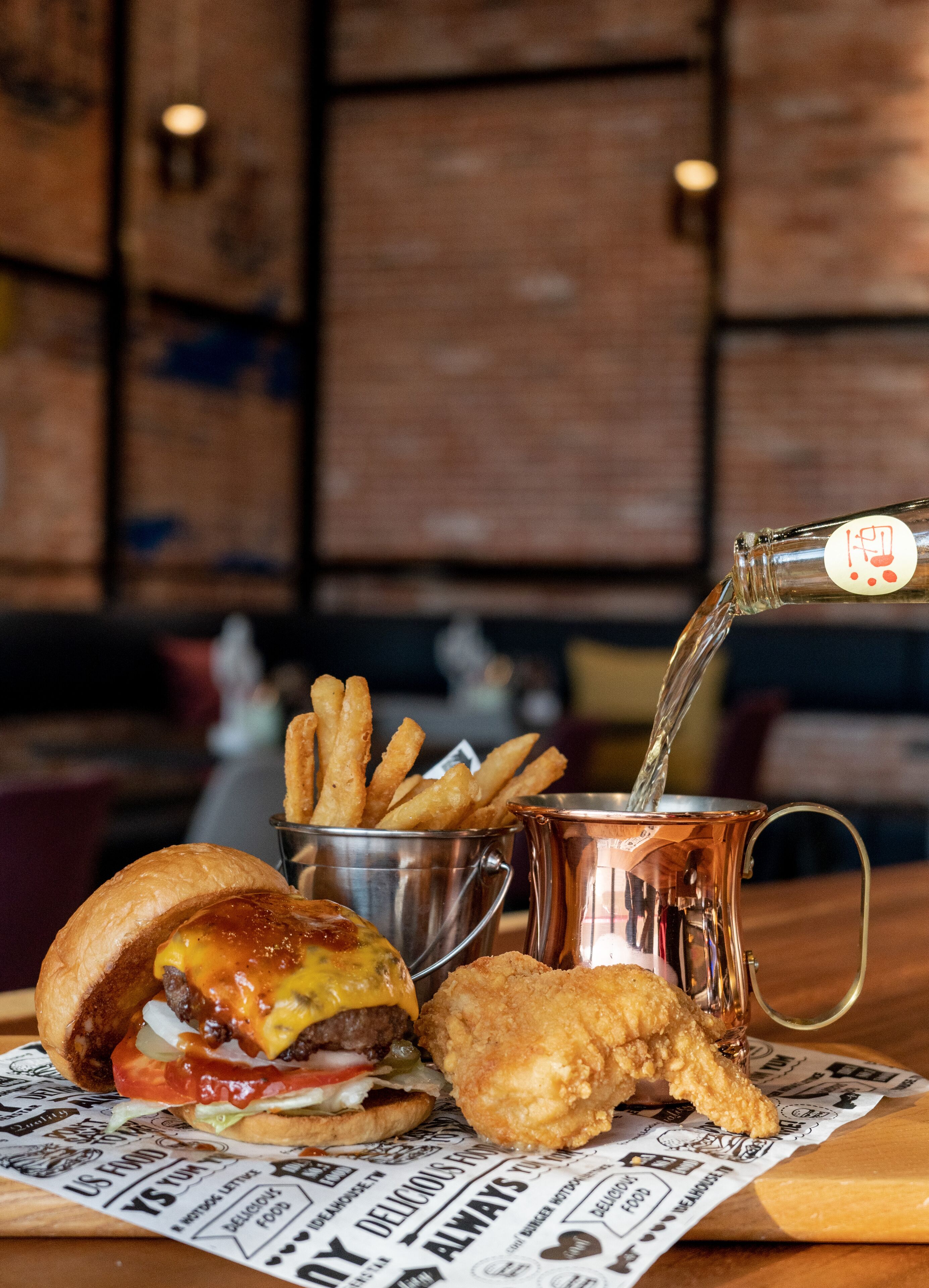 Un repas composé d’un hamburger, de frites et d’un morceau de poulet frit, servi sur une table en bois avec un journal en dessous.
