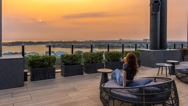 Una mujer sentada en una silla de mimbre contemplando un paisaje urbano al atardecer.