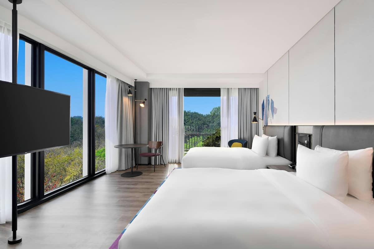 Quarto de hotel moderno com duas camas grandes, televisão de tela plana, mesa de jantar e vista para o exterior.
