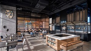 Un comedor moderno con mesas y sillas, una isla central con asientos y una zona de cocina con un letrero que dice 'PICK UP HERE'.