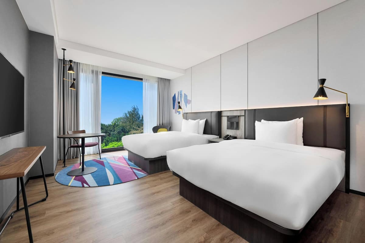 Quarto de hotel moderno com duas camas, uma escrivaninha e vista para o exterior.