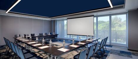 Un salón de conferencias con una gran mesa, sillas, una pantalla de proyector y vistas al exterior con zonas verdes.
