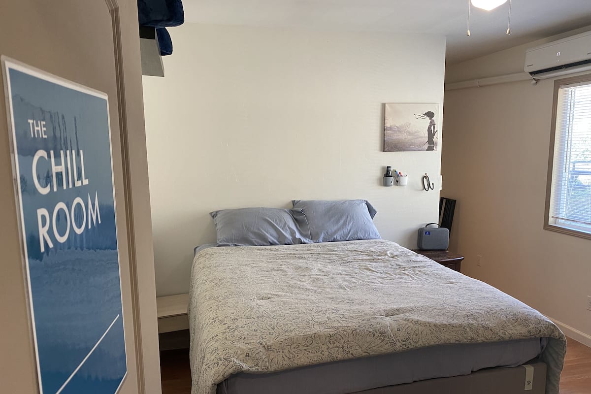 A bedroom with a bed, a bedside table, a wall-mounted air conditioner, a window with blinds, and a poster on the wall.