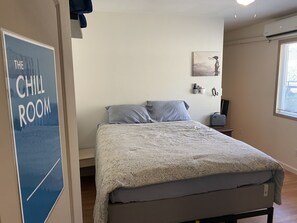 A bedroom with a bed, a bedside table, a wall-mounted air conditioner, a window with blinds, and a poster on the wall. - WHOLE Family - Barrette - Hostel (Sacramento)