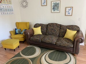 A living room with a brown leather sofa, a yellow armchair, and a matching ottoman. There are two framed artworks on the wall and a decorative wall hanging. - WHOLE Family - Barrette - Hostel (Sacramento)