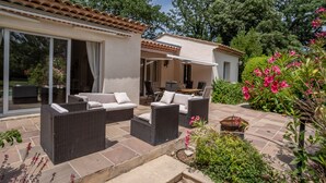 Eine Terrasse mit Korbmöbeln, eine Glastür zu einem Zimmer und ein Garten mit rosa Blumen.