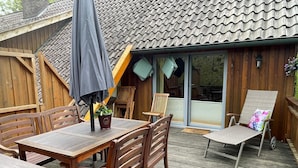 A wooden deck with a table and chairs, a covered umbrella, and a sliding glass door leading to a covered patio area. - Varus vacation apartment in the Kalkriese recreation area surrounded by meadows and forests (Ostercappeln)