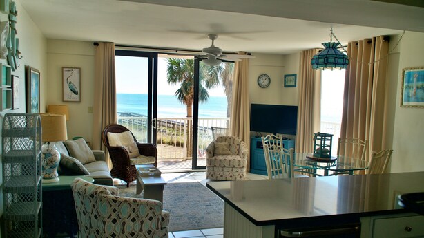 A spacious living room with a large window overlooking the beach, a ceiling fan, and a dining area with a glass table. - Gulf Front Condo, outdoor/ indoor pools, Large wrap around balcony, 3br/2ba (Orange Beach)