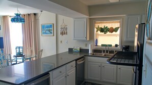 A kitchen with white cabinets, a dishwasher, a sink, and a refrigerator. There is a window with a starfish decoration and a painting on the wall. - Gulf Front Condo, outdoor/ indoor pools, Large wrap around balcony, 3br/2ba (Orange Beach)