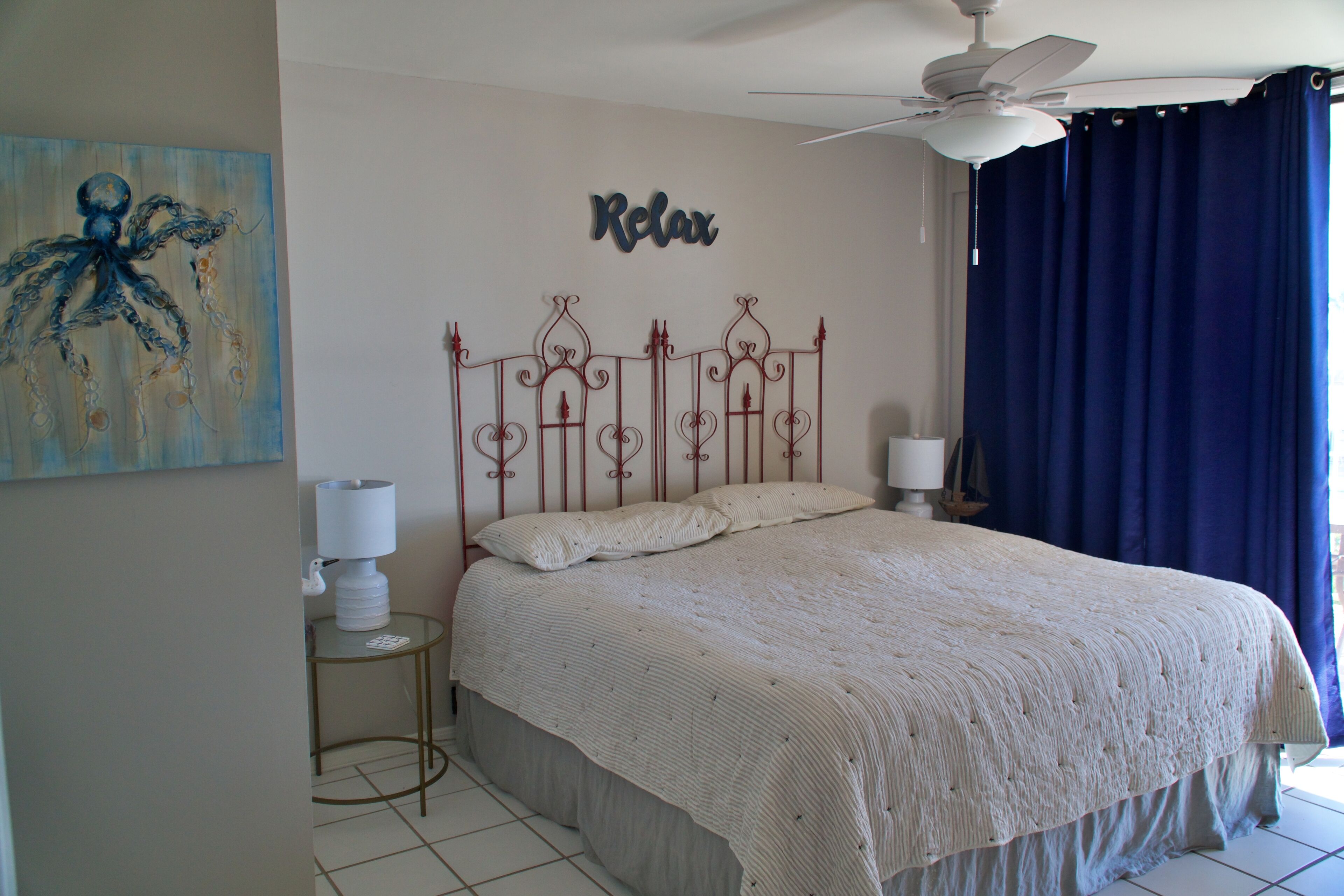 A bedroom with a bed, a ceiling fan, a wall art piece, and a window with curtains.
