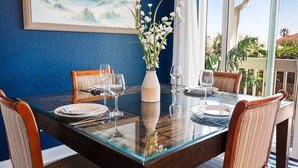 Een eettafel gedekt voor vier personen, met een bloemenvaas met witte bloemen, een schilderij van een strand en uitzicht op palmbomen door een raam.