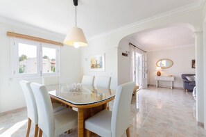 Un coin repas avec une table en verre et des chaises blanches, une lampe suspendue et une vue par la fenêtre.