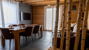 A dining area with a wooden table and chairs, a flat-screen TV mounted on the wall, and a large window with curtains. - Les Jardins du Herisson 4 (BONLIEU)