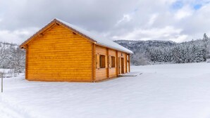 A wooden cabin with a snow-covered roof, situated in a snowy landscape with trees and hills in the background. - Les Jardins du Herisson 4 (BONLIEU)