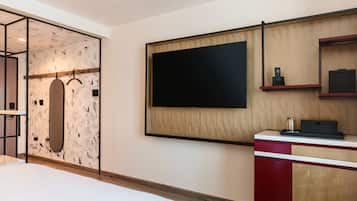 A modern hotel room with a large bed, a wooden wardrobe, a flat-screen TV mounted on the wall, and a small desk with a telephone and a tray.