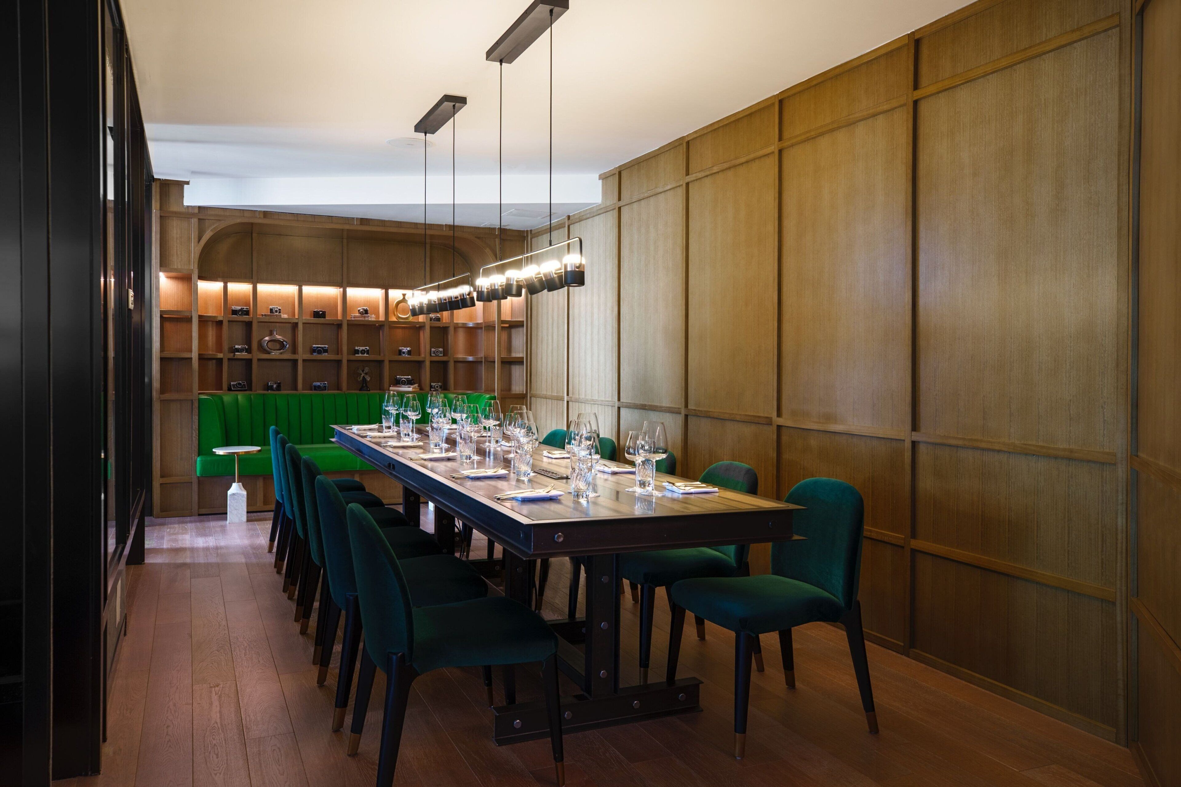 A dining area with a long table set for a meal, surrounded by chairs, with a wooden wall and shelves in the background.