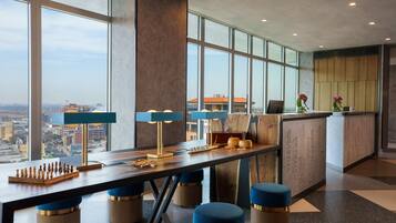 Een moderne hotel lobby met een groot raam met uitzicht op de stad, een receptiebalie en een schaakbord op de tafel.