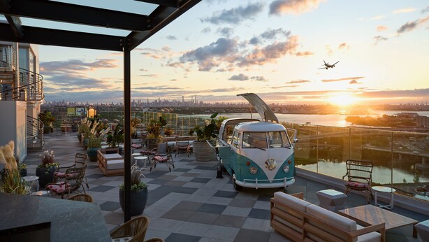 Terraza en la azotea con una furgoneta camper VW de estilo vintage, mobiliario exterior y vistas a un horizonte urbano al atardecer.
