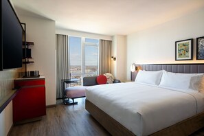 A hotel room with a large bed, a desk, a chair, a television, and a window with a city view.