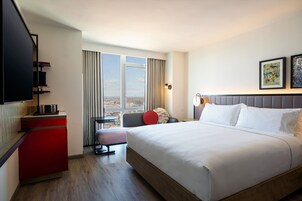A hotel room with a large bed, a desk, a chair, a television, and a window with a city view.