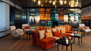 Un área de descanso moderna con un sofá grande naranja, varios sillones y una mesa de centro. Se observan luces de techo y una cocina al fondo.