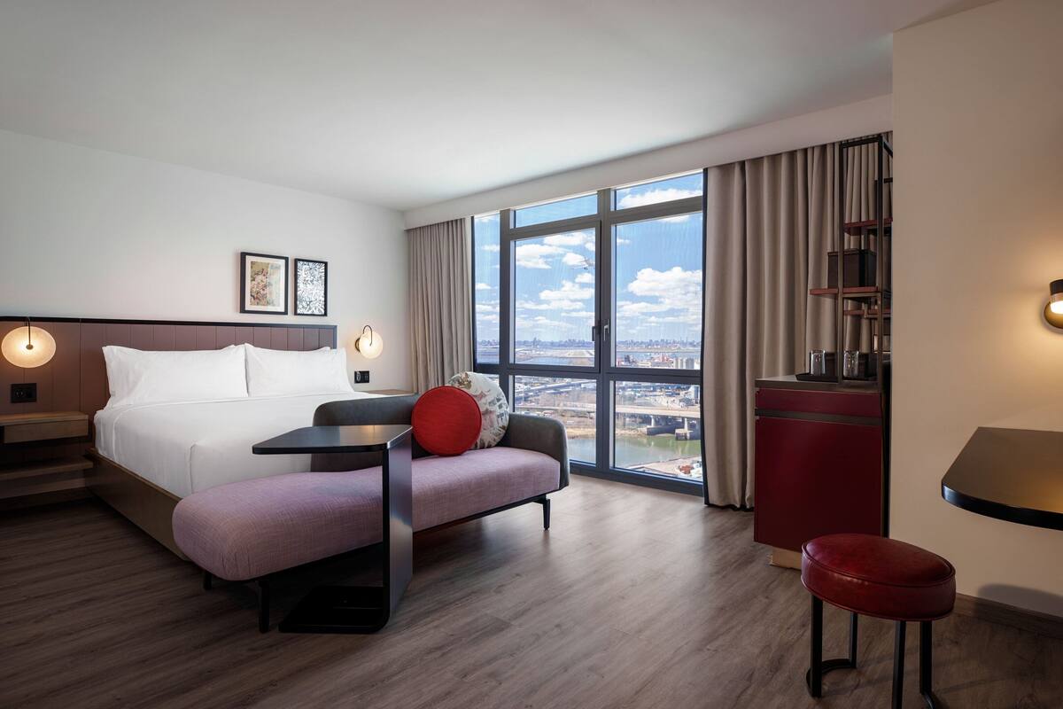 Habitación de hotel moderna con una cama grande, un escritorio, una silla roja y vistas de la ciudad a través de amplios ventanales.