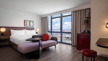 Habitación de hotel moderna con una cama grande, un escritorio, una silla roja y vistas de la ciudad a través de amplios ventanales.
