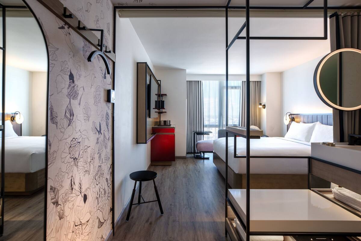 Una habitación de hotel moderna con una cama, un escritorio, una silla, un taburete pequeño, un armario rojo, un espejo y una ventana con cortinas.
