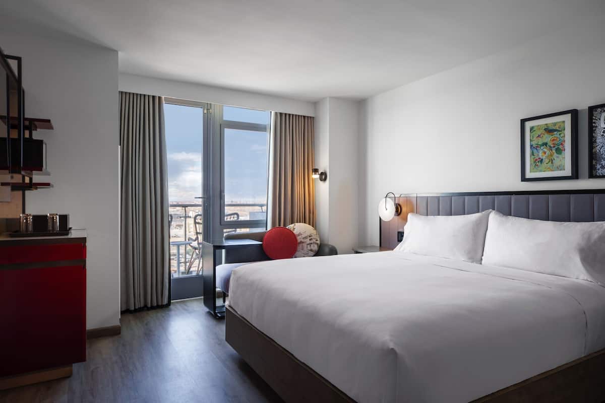 Habitación de hotel con una cama grande, un escritorio con computadora, una silla y vista a la ciudad desde la ventana.