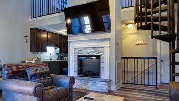 A spacious living room with a fireplace, a mounted TV, and a staircase leading to an upper level.